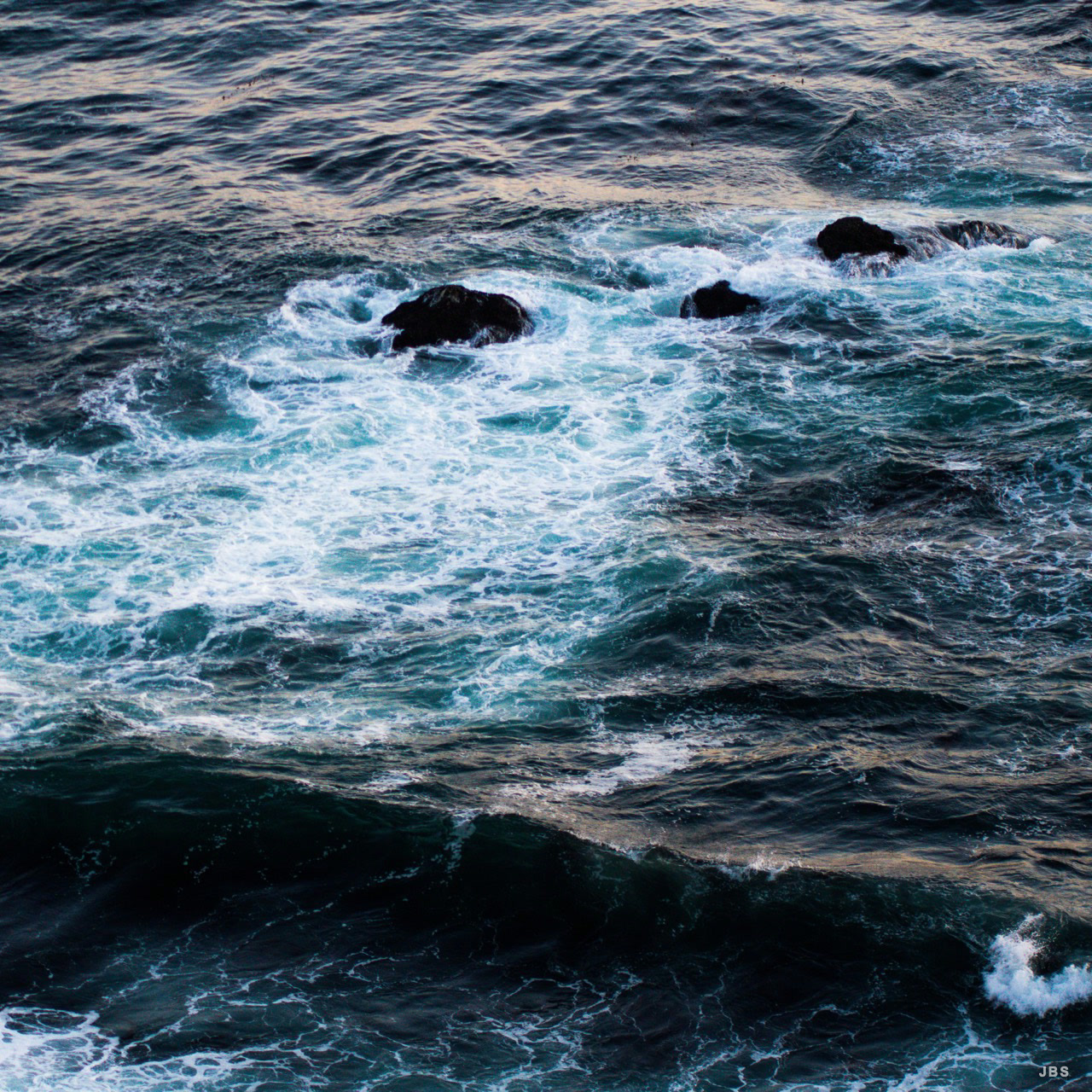Dark ocean swells breaking into turquoise foam around black rocks