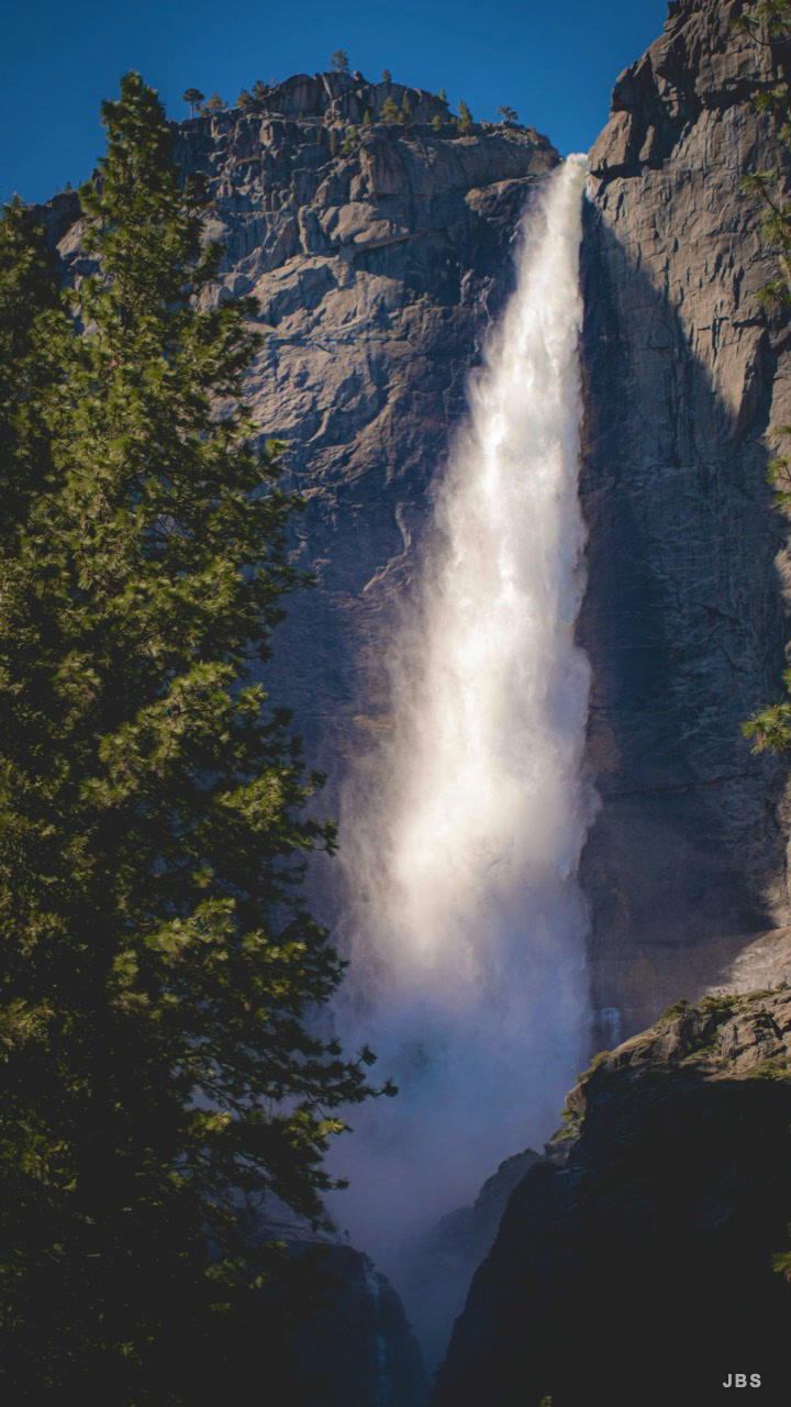 A tall waterfall illuminated against a granite wall and dark pines